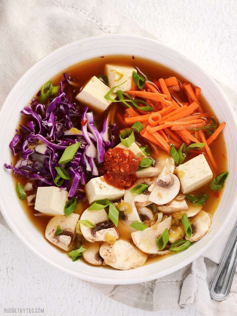 Overhead view of a bowl full of hot and sour soup with vegetables and tofu. Garnished with a dab of chili garlic sauce.