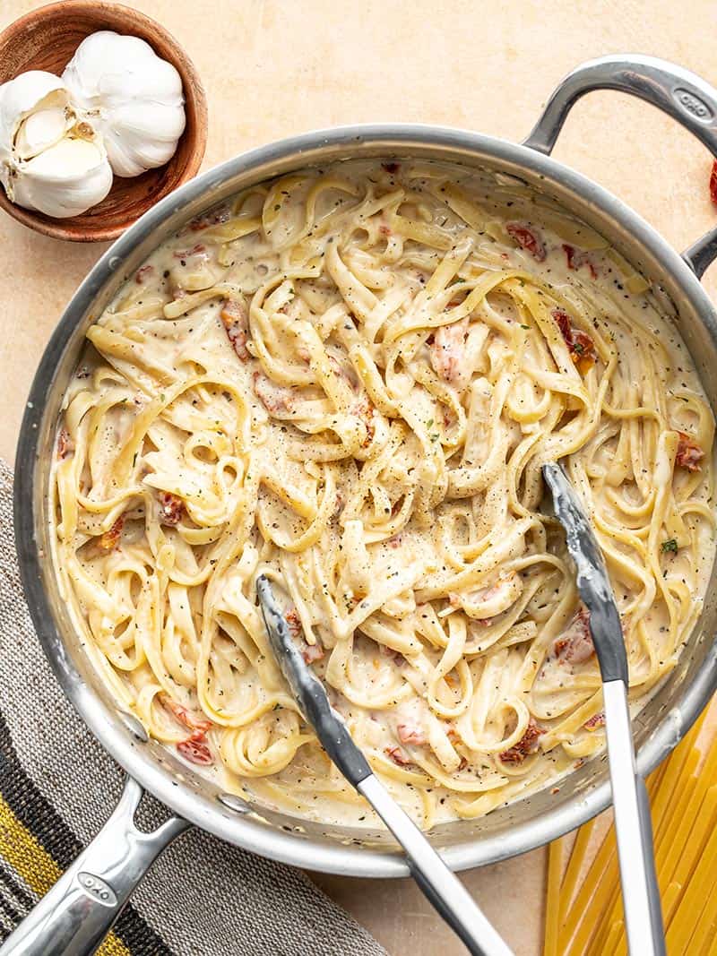 Creamy Sun Dried Tomato Pasta in a deep skillet with tongs, a bowl with garlic and dry pasta on the side