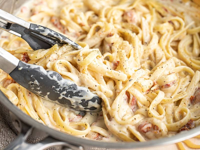 Close up of tongs picking up a clump of creamy pasta from the skillet