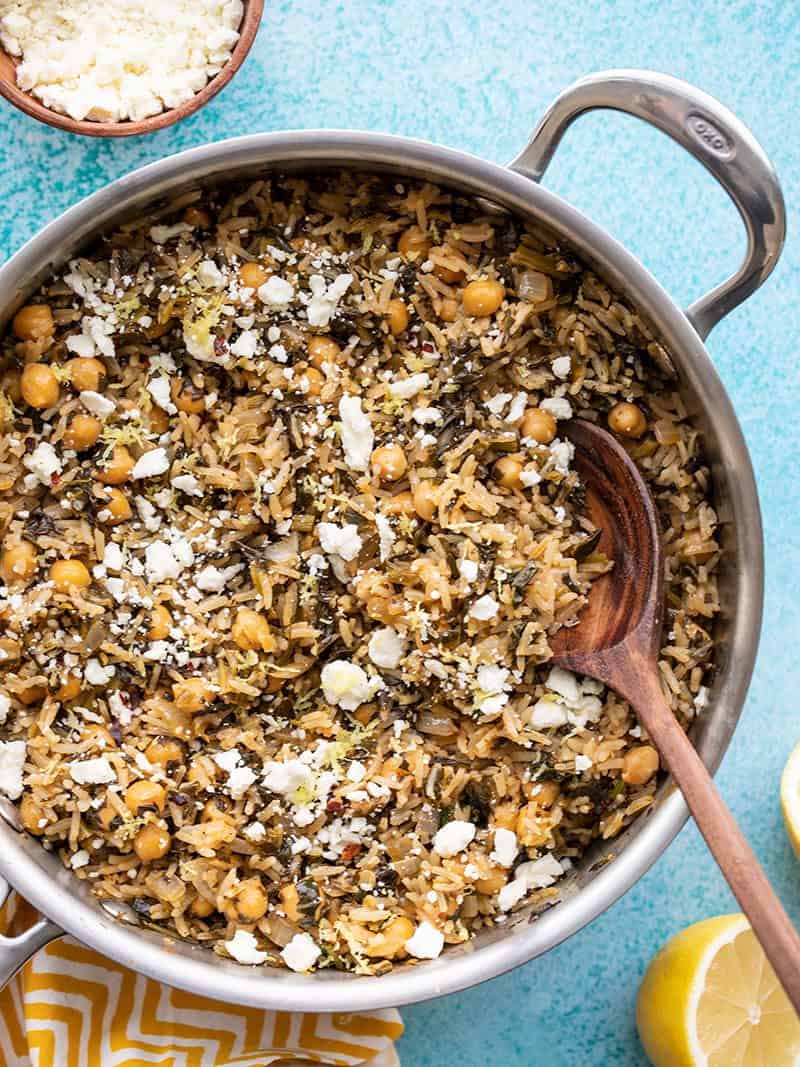 Close up overhead view of a skillet full of Spinach and Chickpea Rice Pilaf with a wooden spoon and bowl of feta on the side.