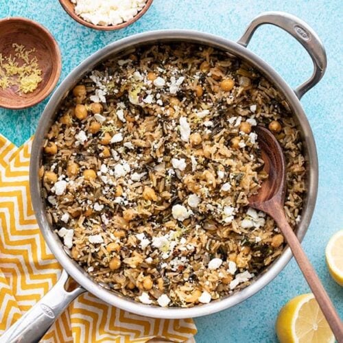 Skillet full of Spinach and Chickpea Rice Pilaf next to a cut lemon and small dish of feta, on a blue background