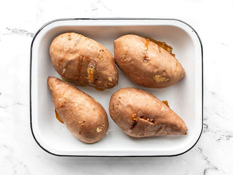 Baked sweet potatoes in the baking dish with some juices bubbling out of the fork holes