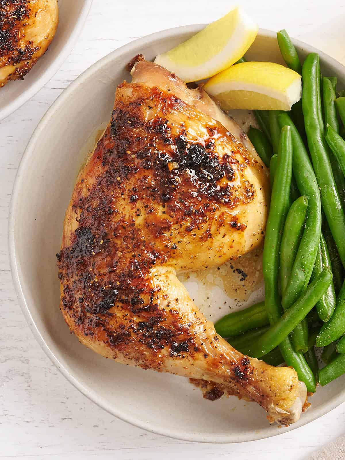 Overhead view of a baked chicken leg on a plate with green beans and lemon wedges.