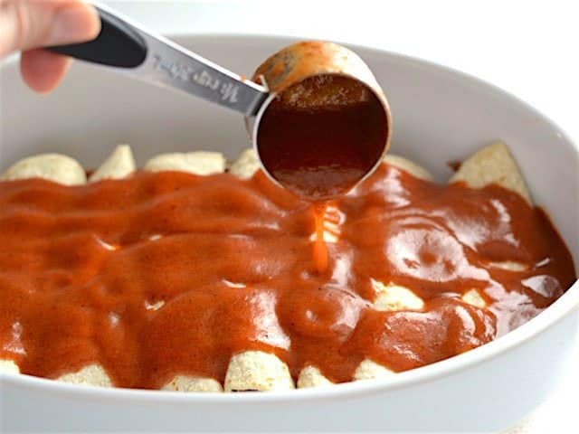 Enchilada sauce being scooped over the casserole dish of enchiladas