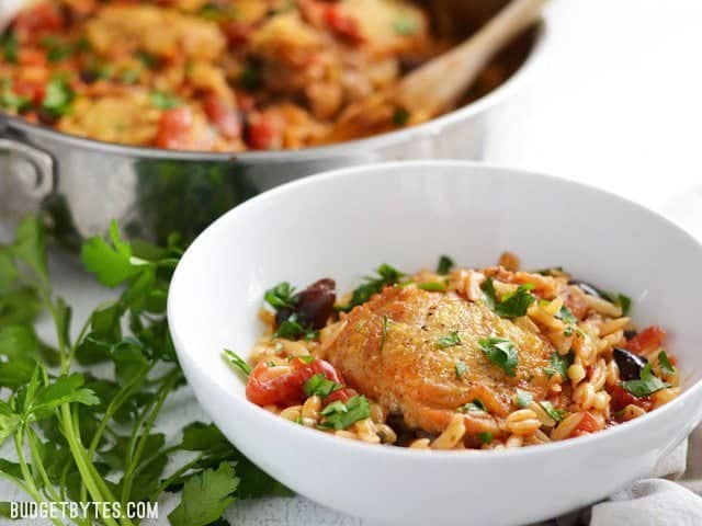 A bowl of chicken with orzo and olives, the skillet in the background