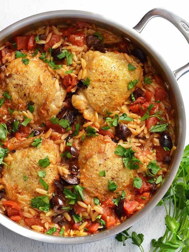 Overhead view of a skillet full of chicken and orzo with olives and tomatoes, parsley on the side