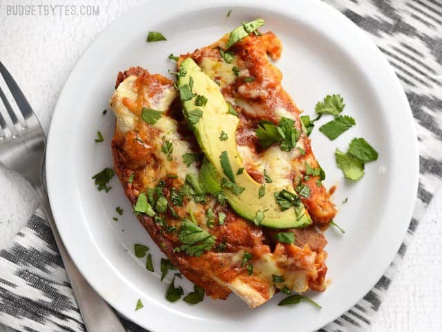 Two baked enchiladas on a plate, topped with an avocado slice and chopped cilantro.