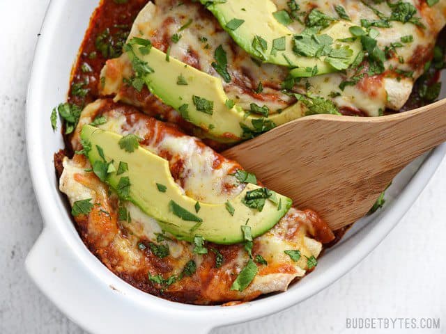 These easy Bean and Cheese Enchiladas are anything but boring. A few extra details take them from a simple to spectacular weeknight dinner. BudgetBytes.com A wooden spatula lifting two cheese enchiladas out of a casserole dish