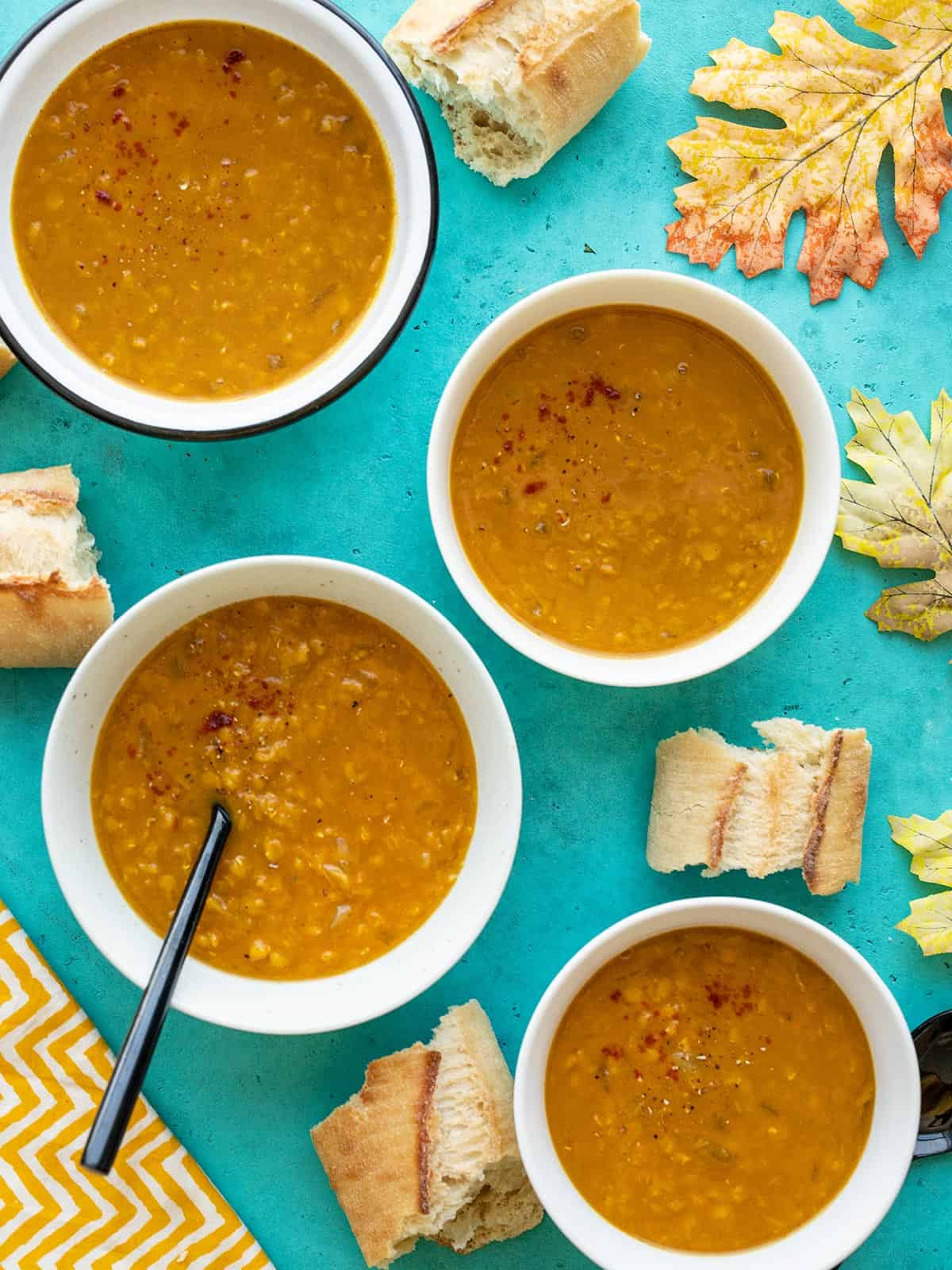 Four bowls of curried red lentil and pumpkin soup with bread and leaves scattered around the bowls