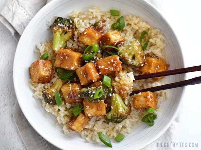 Chopsticks digging into a bowl of Pan Fried Sesame Tofu with Broccoli and brown rice. 