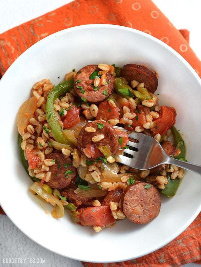 A bowl full of smoked sausage with peppers, onions, and farro. A fork pierced through a piece of sausage in the center