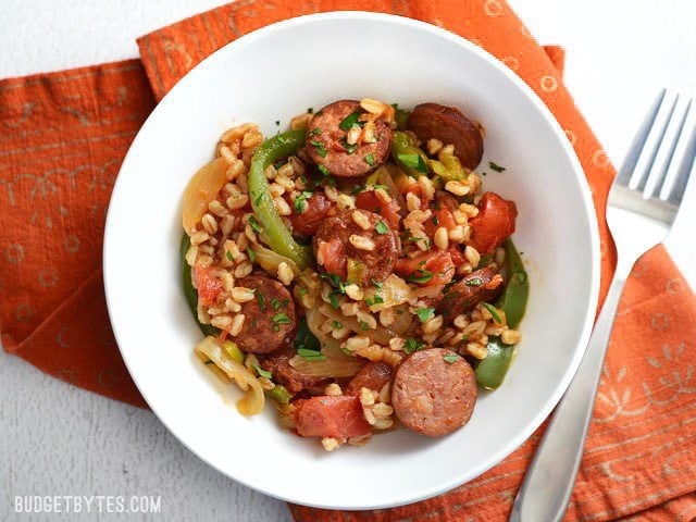 Smoked sausage and peppers with farro served in a bowl, on an orange napkin, fork on the side