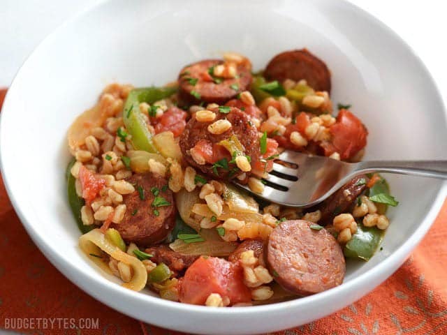 Side view of a bowl full of smokes sausage with peppers, onions, and farro. A fork in the middle