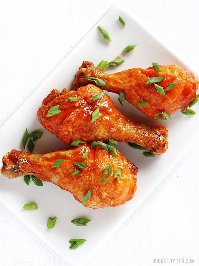 Three honey sriracha baked chicken drumsticks on a white rectangular plate, garnished with sliced green onion.