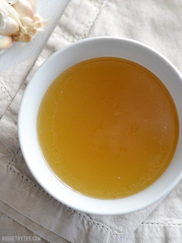 Overhead view of a bowl of golden chicken stock, on a cream colored napkin, a bulb of garlic on the side.