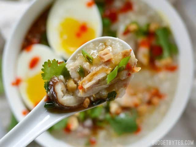 Close up of a spoonful of congee with toppings, the bowl in the background