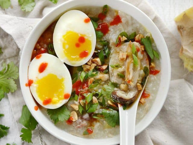 A bowl of congee with toppings, including a soft boiled egg and drizzle of sriracha, a spoon in the side