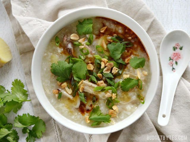 A white bowl full of congee, topped with cilantro, peanuts, soy sauce, and sesame oil