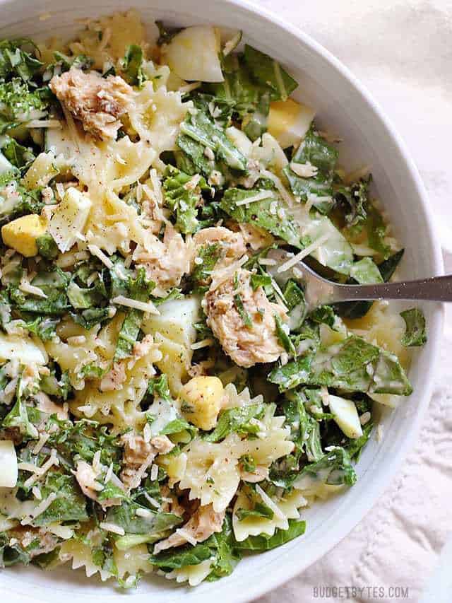 Overhead view of a white bowl full of kale and salmon caesar salad, a fork stuck in the middle.