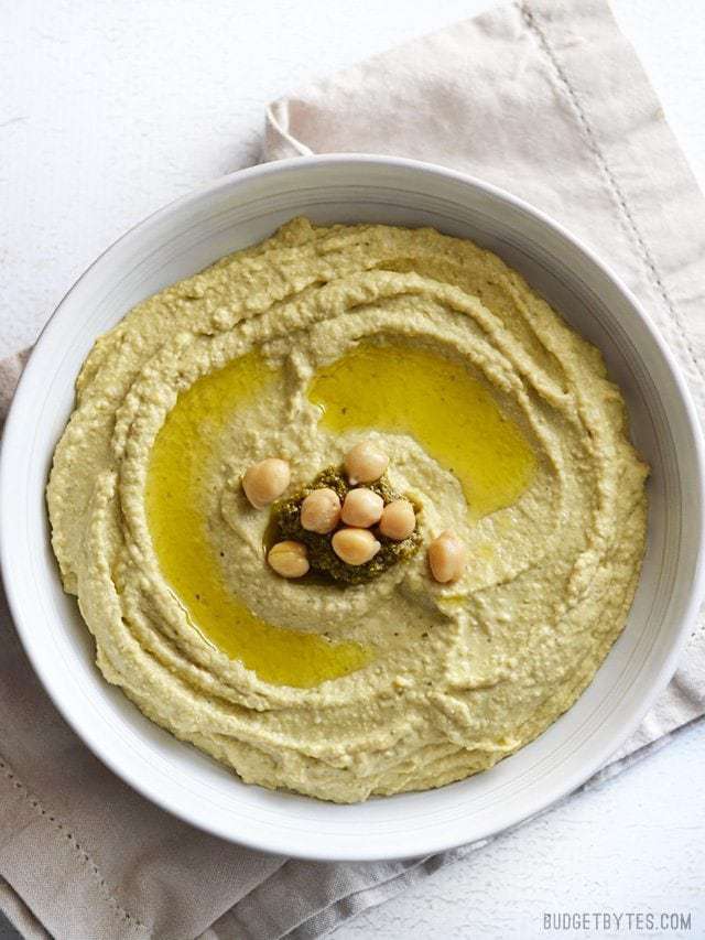 Overhead view of a bowl full of pesto hummus garnished with olive oil, a dollop of pesto, and a few whole chickpeas