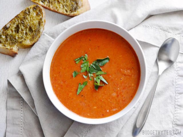 Overhead view of a bowl of Roasted Red Pepper and Tomato Soup garnished with parsley, pesto toast on the side