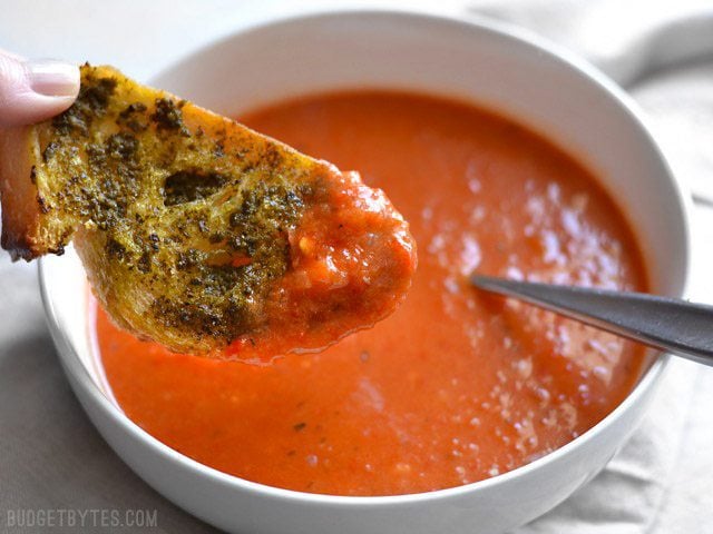 A piece of pesto toast dipped into a bowl of roasted red pepper and tomato soup.