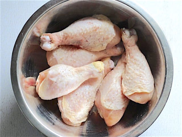 Raw chicken drumsticks in a metal mixing bowl, seasoned with a pinch of salt and pepper