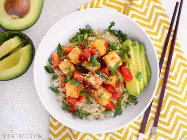 Overhead view of a Sweet Chili Tofu Bowl on a yellow and white napkin, a cut open avocado on the side