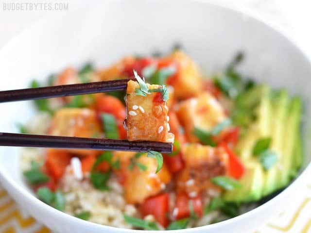 Close up of chopsticks holding one tofu cube coated in sweet chili sauce, the bowl in the background