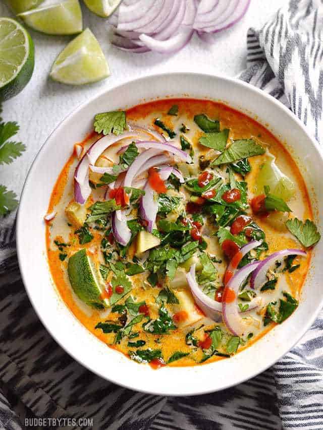 Overhead view of a bowl of Thai Curry Vegetable Soup garnished with lime wedges, sliced red onion, fresh cilantro, and a drizzle of sriracha