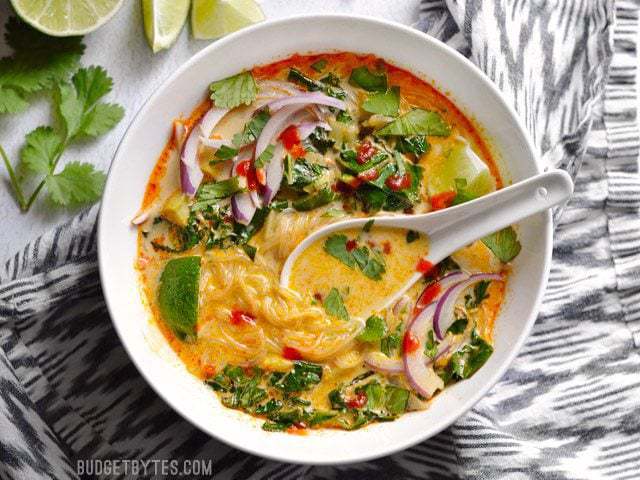 Overhead view of a bowl of Thai Curry Vegetable Soup with a ceramic spoon, sitting on a striped napkin with lime wedges and cilantro on the side.