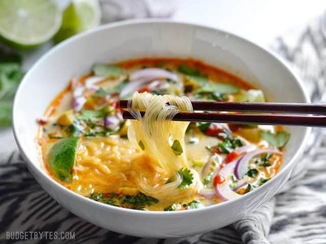 Noodles being lifted out of a bowl of Thai Curry Vegetable Soup with a pair of wooden chopsticks