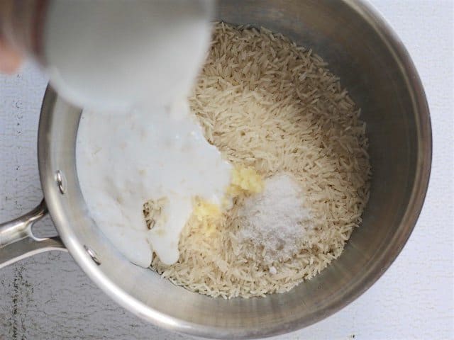 Pouring coconut milk into a sauce pot of rice with minced garlic and salt to make the Coconut Rice