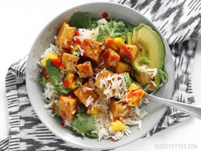 A fork digging into a Mango Coconut Tofu Bowl, with sriracha drizzled over top