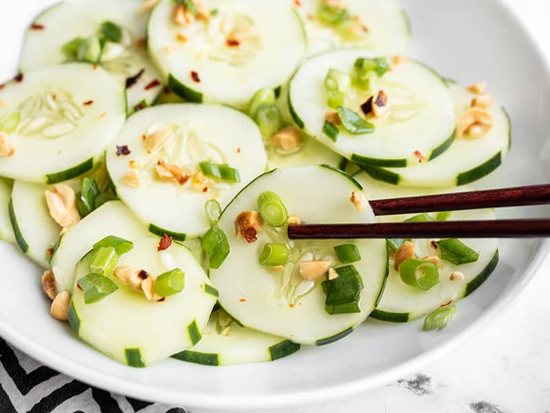Side view of a bowl of sesame cucumber salad, chopsticks picking up one slice of cucumber