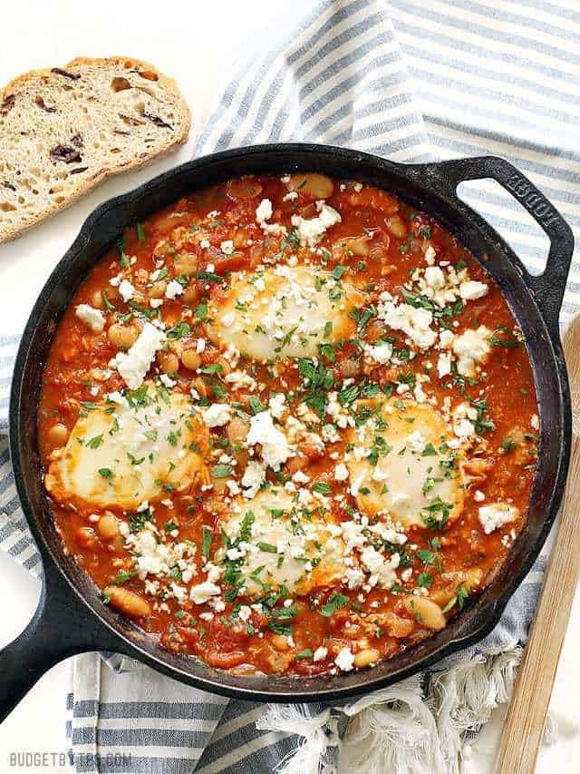 A cast iron skillet full of Smoky White Bean Shakshuka topped with chopped parsley, a slice of bread on the side.