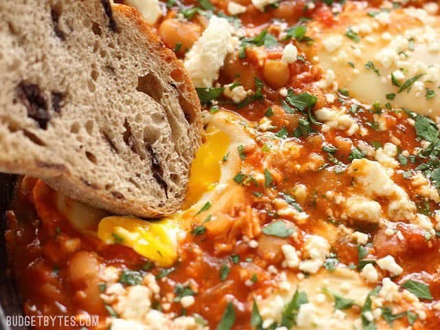 Close up of a slice of bread being dipped into a runny. yolk in the Smoky White Bean Shakshuka 