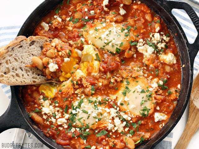 A piece of bread dipping into a runny yolk in a skillet full of Smoky White Bean Shakshuka 