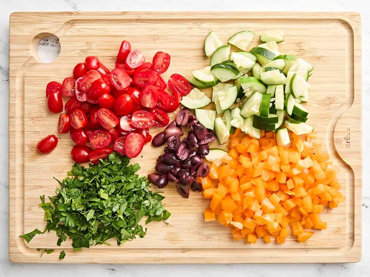 Diced cucumber, bell peppers, tomatoes, and parsley leaves on a wooden cutting board.