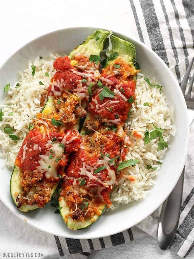 Two Italian Sausage Stuffed Zucchini boats in a bowl over a bed of rice with a striped napkin in back