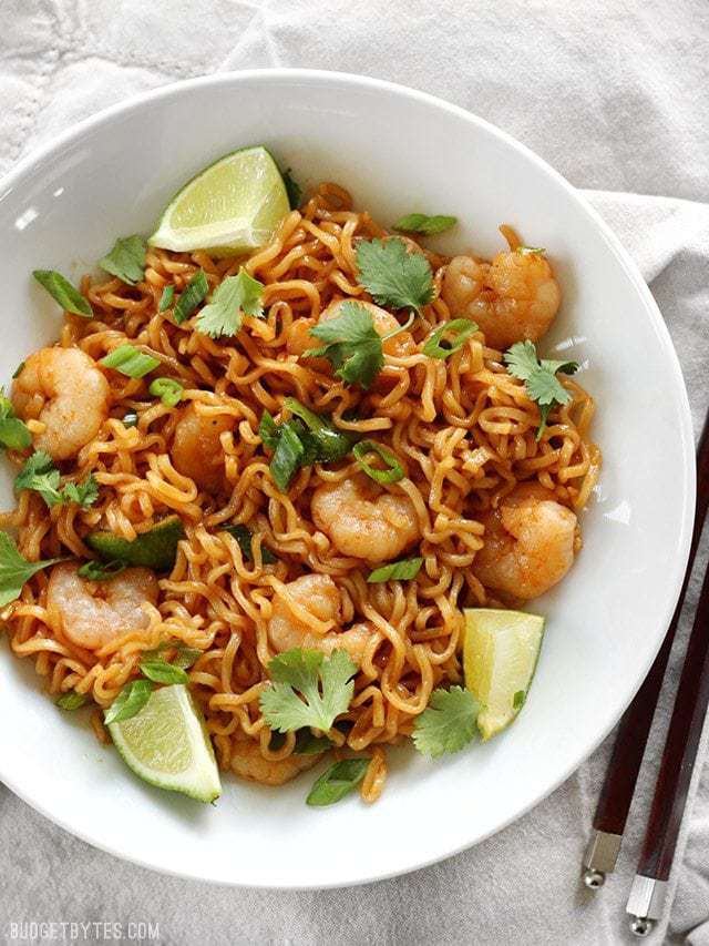 Overhead view of a bowl full of Lime Shrimp Dragon Noodles garnished with lime wedges and fresh cilantro. Chopsticks on the side.