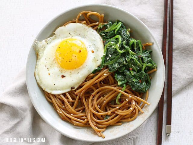 Overhead view of a bowl full of Sesame Noodles with Wilted Greens topped with a fried egg and chopsticks on the side