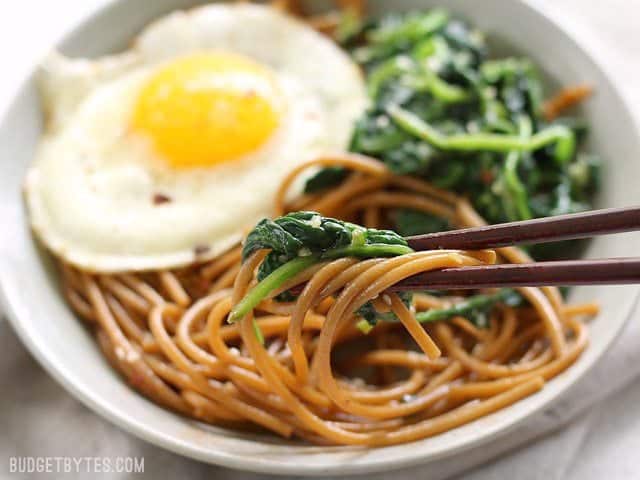 Close up side view of a bowl of Sesame Noodles with Wilted Greens with chopsticks picking up some noodles