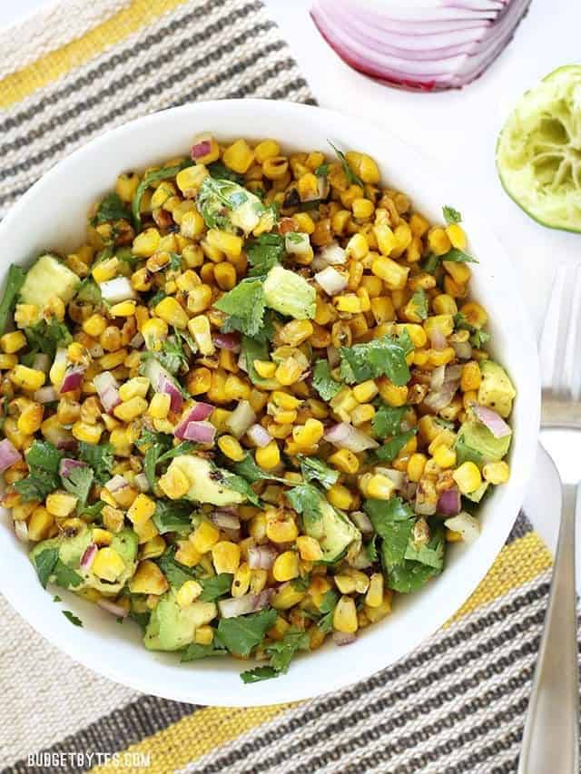 Overhead view of a bowl full of Warm Corn and Avocado Salad on a striped placemat with red onion and lime on the side