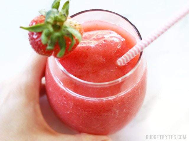 A hand holding a glass of Strawberry Rosé Slush with a pink paper straw and fresh strawberry garnish