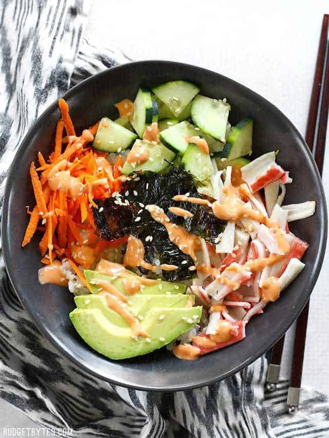 Overhead view of a homemade sushi bowl with krab, avocado, cucumber, carrot, seaweed, and sriracha mayo in a black bowl