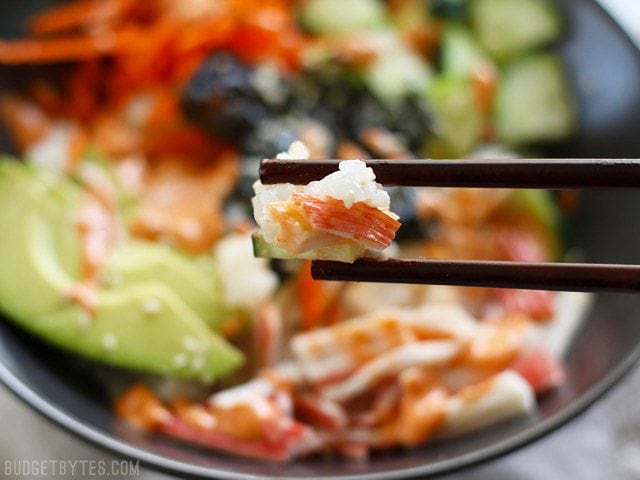 Close up of chopsticks holding a chunk of sushi rice and crab stick, with sushi bowls in the background.