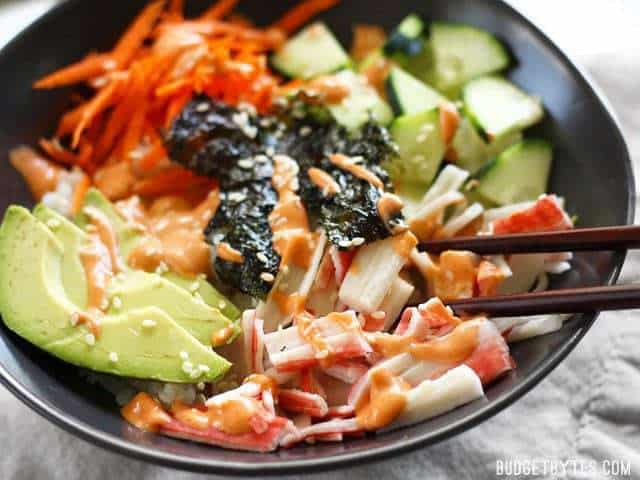 close up side view of a homemade sushi bowl with chopsticks picking up a piece of crab stick