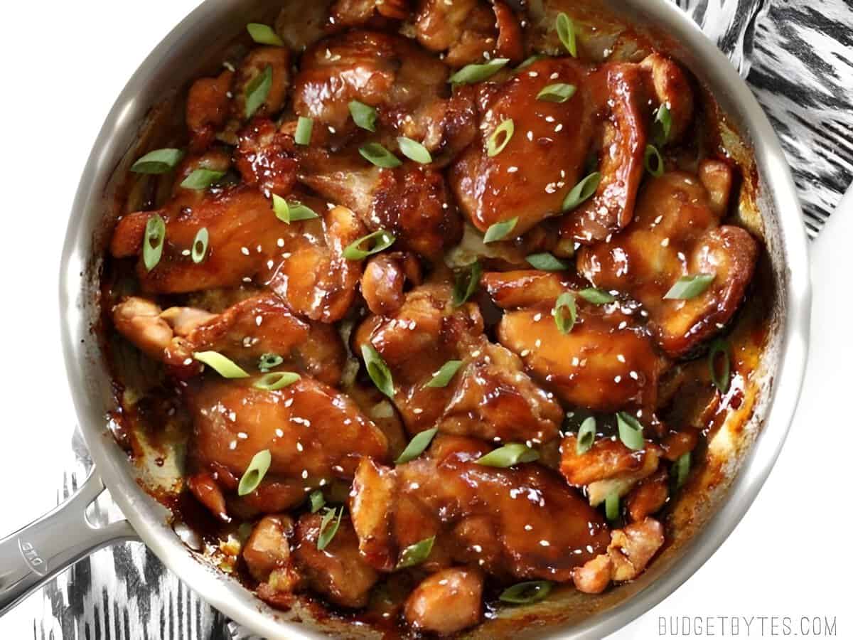 Sticky ginger soy glazed chicken in a skillet with sliced green onions.