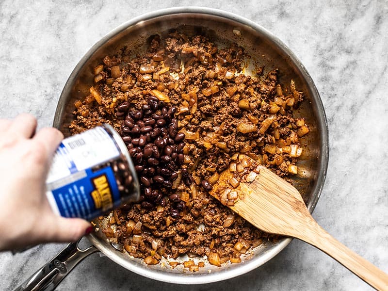 Canned black beans being poured into the skillet with the beef mixture